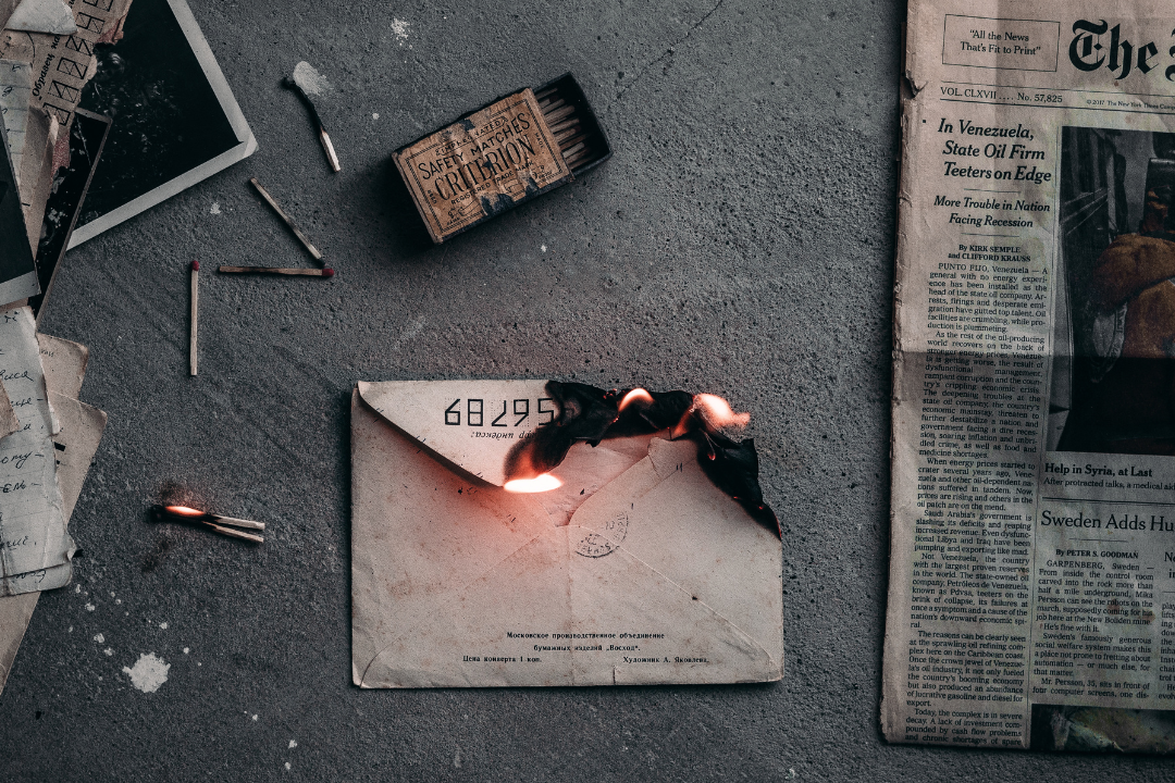 Letter in an envelope that is burning on a table, beside a newspaper, matches, and photographs.