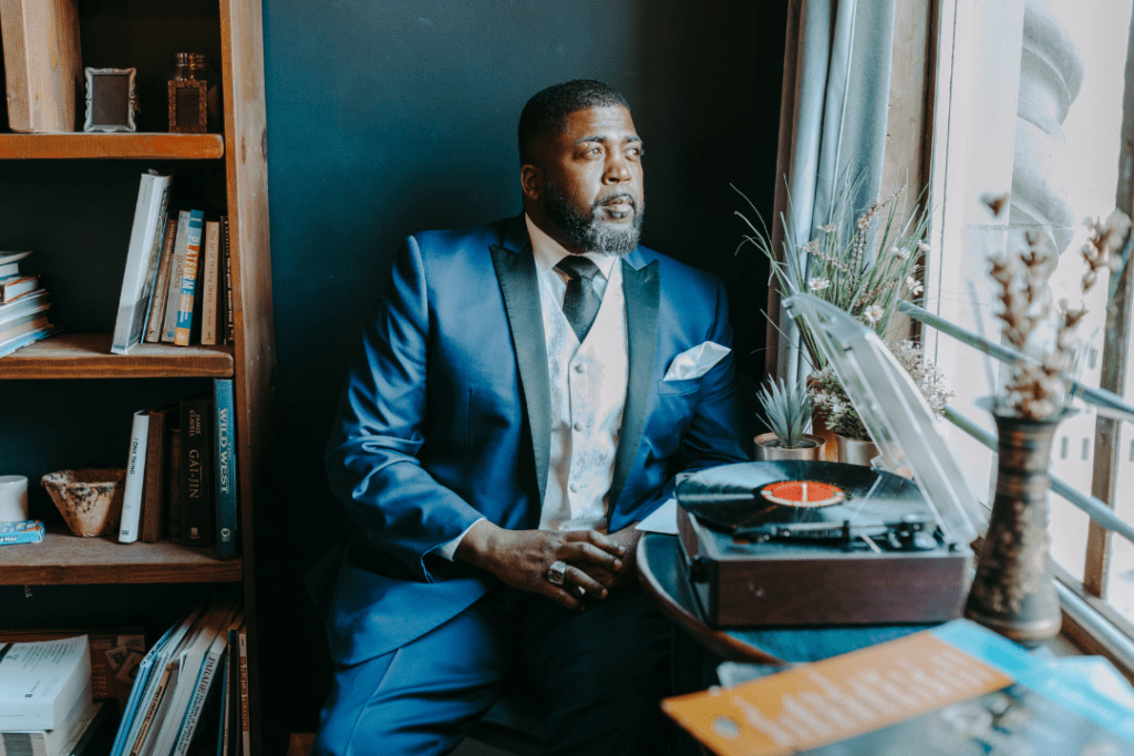 Man wearing a blue suit sitting beside a window and record player. The man is looking out the window.