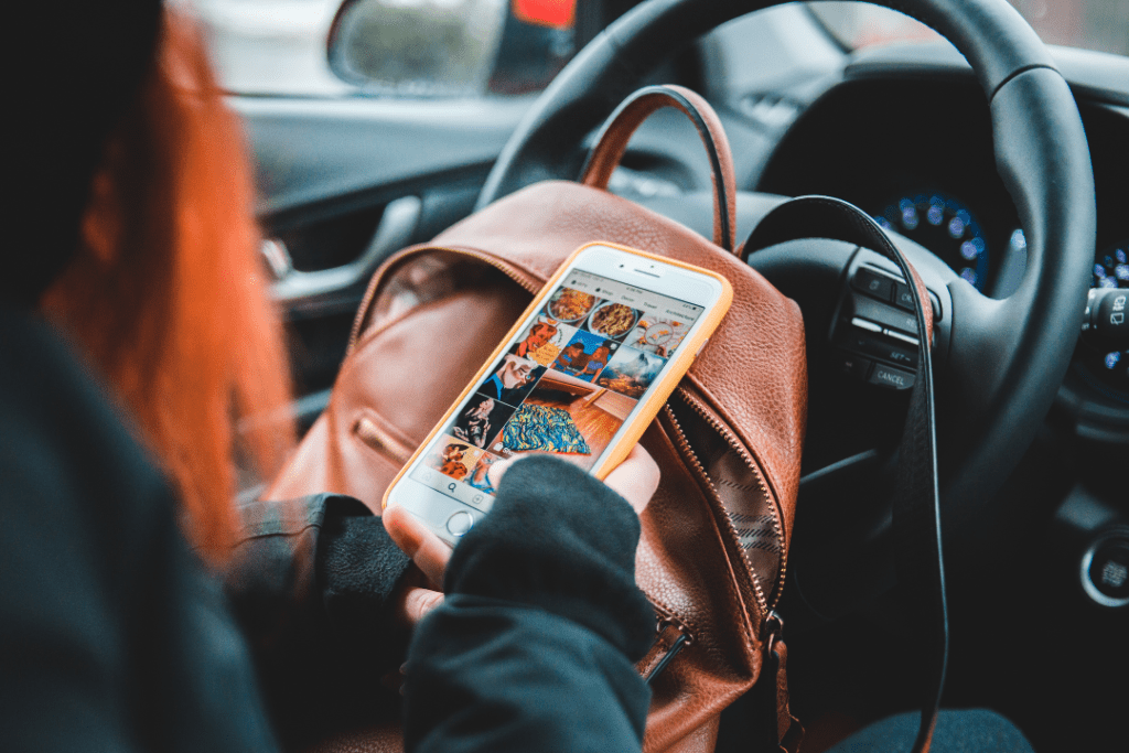 Person with red hair sitting in a car holding and looking at a mobile phone in the drivers seat. They look like they are scrolling on Instagram.