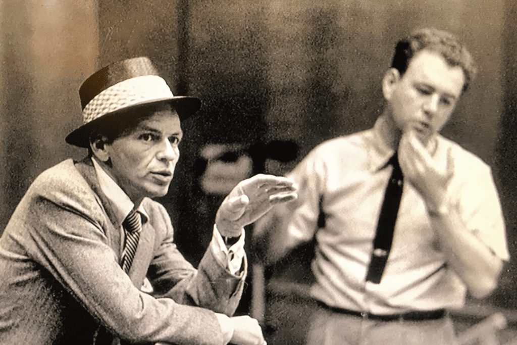 Frank Sinatra with nelson Riddle. One man in the foreground wears a suit and hat, he is talking and gesturing with one hand. The other man is in the background standing, touching his chin.