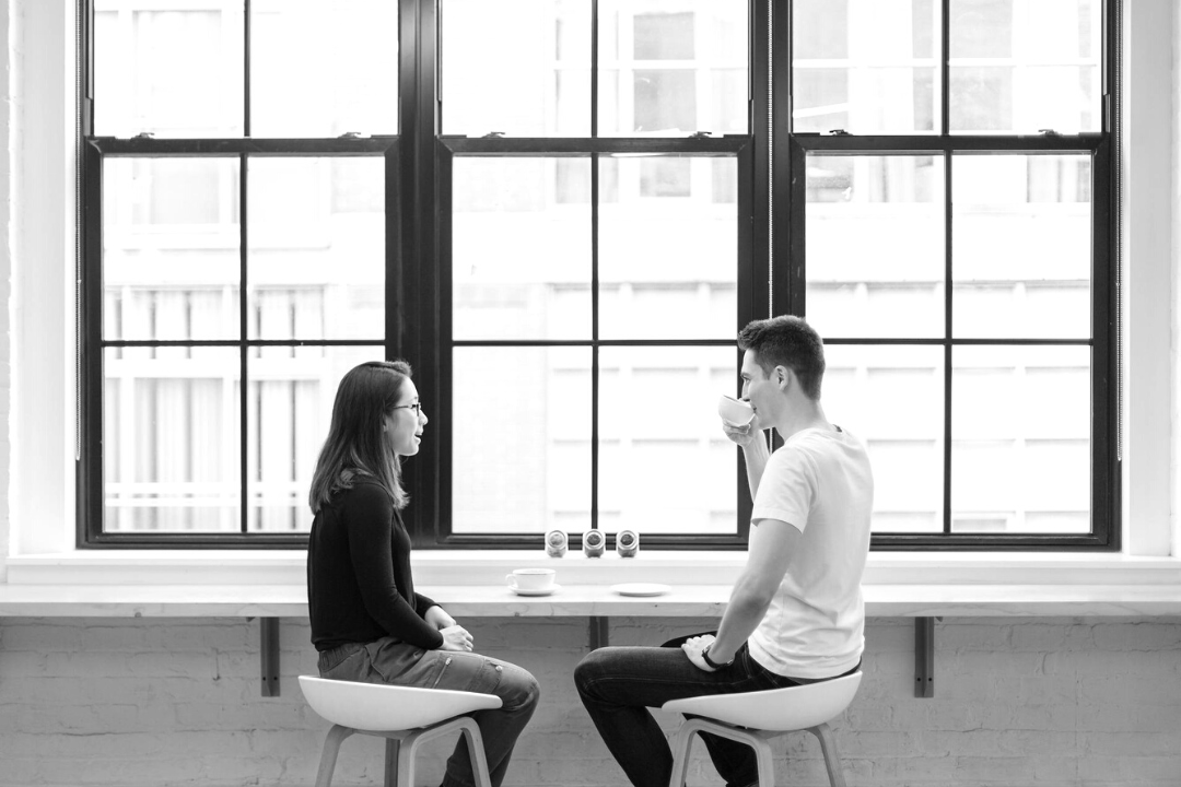 Woman and a man drinking coffee, sitting near large windows.