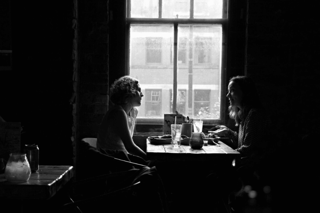 Two women having a meal and serious conversation at a wooden table, sitting near a window.