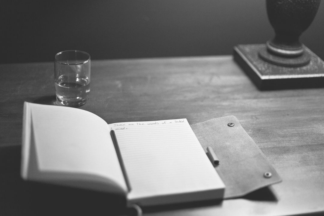 Notebook open on a desk with a glass of water and a vase on the desk.