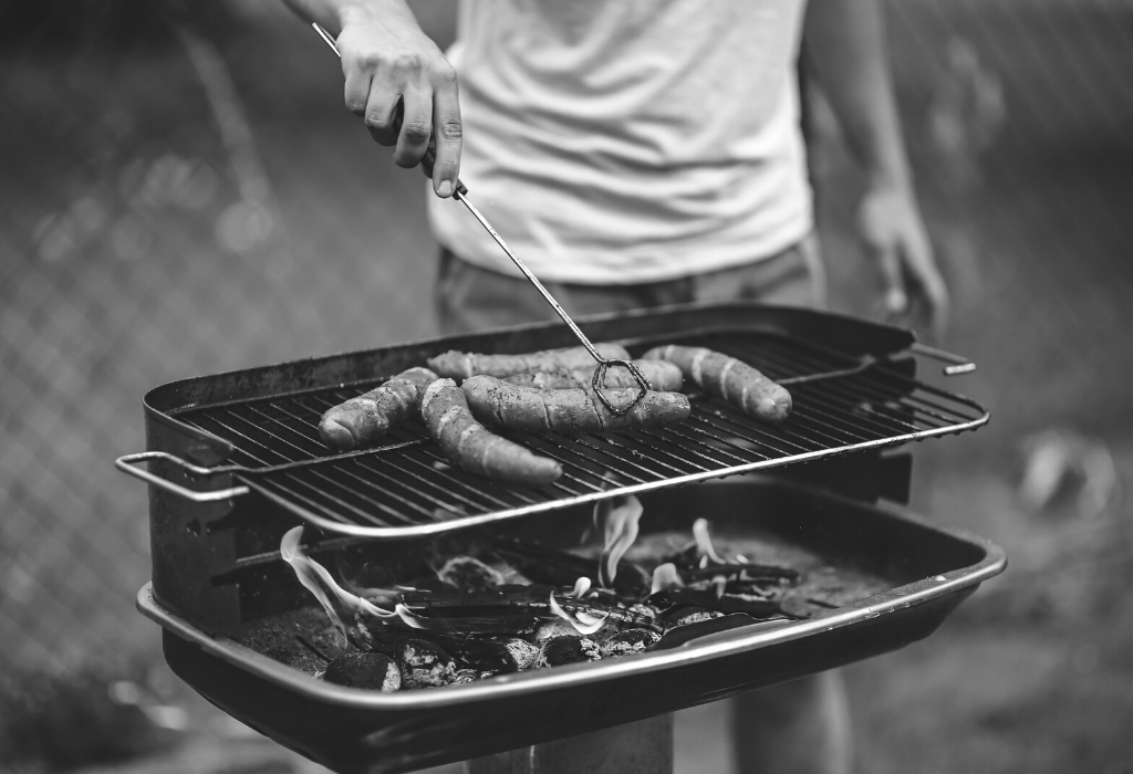 Black and white image of a man in shorts and a tshirt, cooking sausages on a barbecue.