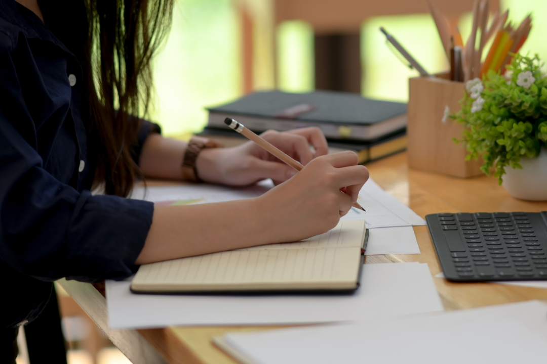 Female with long dark hair sitting at a desk with a notebook and pen. A laptop, notebooks, pencil box and plant are on the desk.