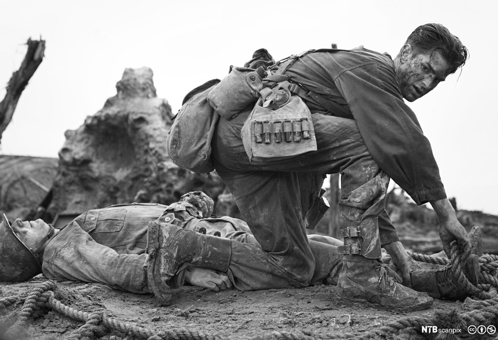 Black and white image of two soldiers. One soldier is injured on the ground and the other soldier is rescuing him by placing a rope around the injured soldier's foot.