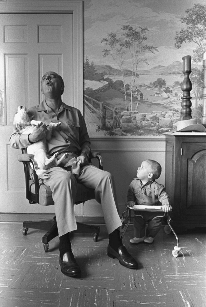 President Lyndon B Johnson sitting on a chair with wheels, holding his dog while they howl. This grandson is a toddler, looking up at him.