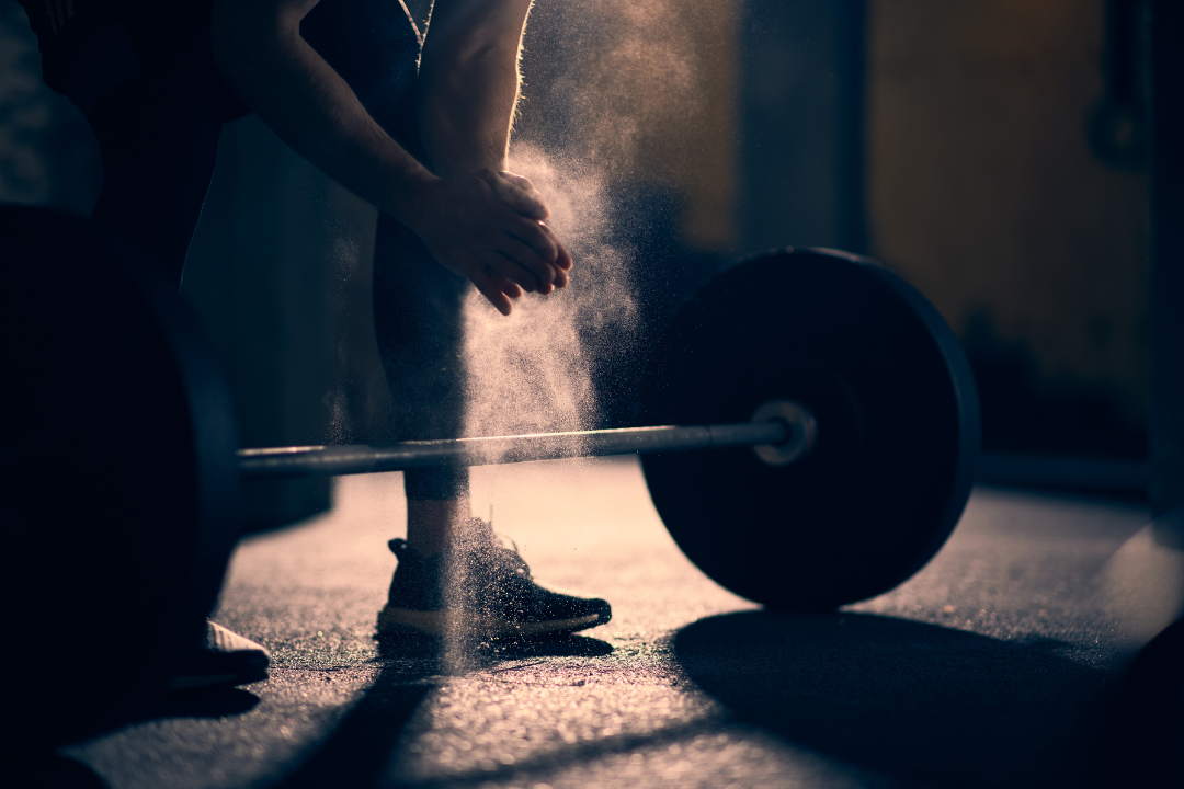 Person wearing runners, crouched down over a barbell, brushing chalk off their hands.